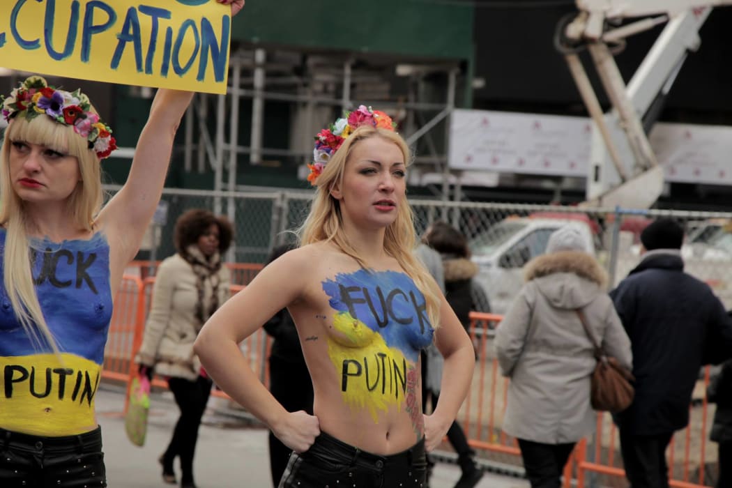a woman standing amid a protest, bare-chested, with body paint saying "fuck putin"