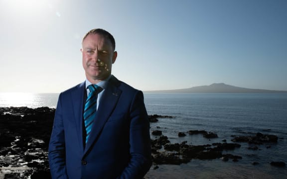 Climate Change minister Simon Watts, wearing a blue suit, stands in bright sunlight near a rocky shore, with Rangitoto Island in the background.