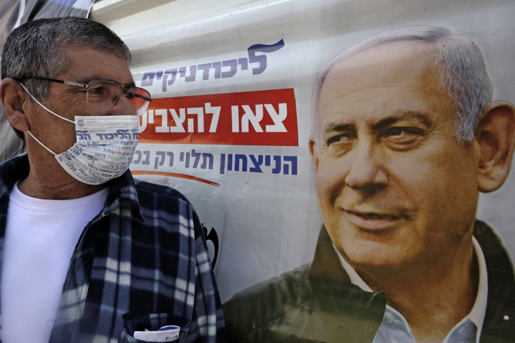 An Israeli man, wearing a mask with the Likud party logo, looks at a poster of Israeli Prime Minister Benjamin Netanyahu, chairperson of the party, in Israel's southern city of Beersheva on March 7, 2021, ahead of the legislative election.