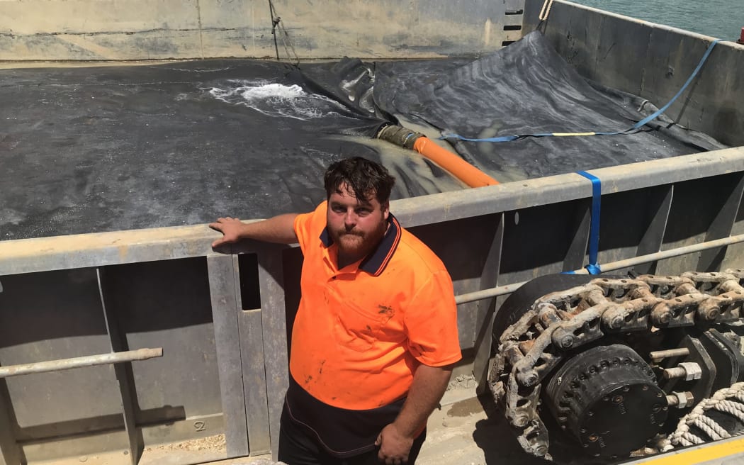 Andrew Johnson with the huge sealed geotech bag on his barge made in Thames slowly starting to fill up with mixed caulerpa, sediment and seawater dredgings from the Omākiwi Cove seafloor. The 1.5m high wall around the bag provides added protection against tiny pieces of caulerpa being spread around as the plants are gently vacuumed up