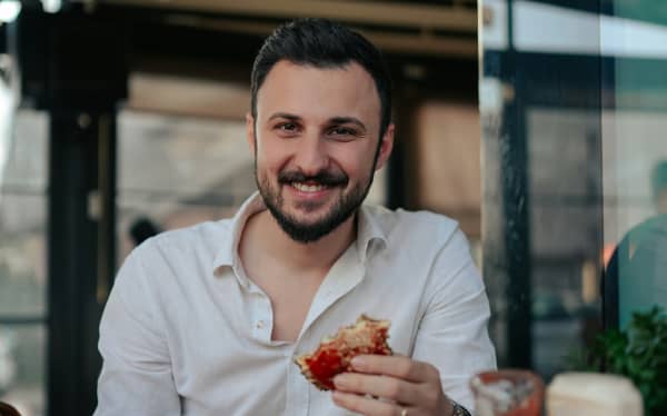 A bearded man in a white shirt smiles as he eats a burger.