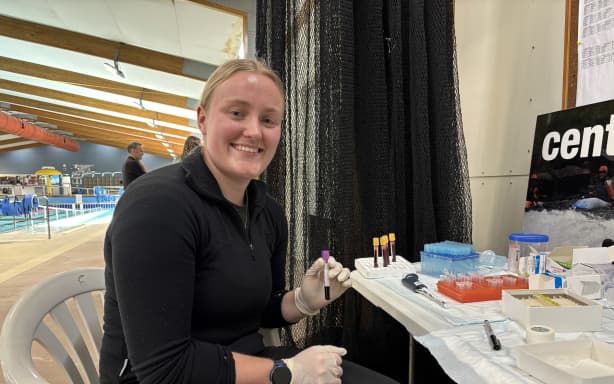 Emma is wearing a black fleece and tracksuit pants, sitting in a white plastic chair at a table. She's holding a plastic vial of blood with a purple cap. On the table are an array of tubes, pipettes, pipette tips, markers and boxes. She is indoors, with a little sliver of Alexandra's indoor pool visible in the background. She is turned toward the camera, smiling, with the vial in her gloved left hand, her right is resting on her lap.