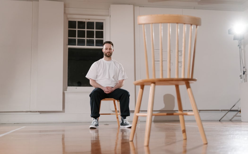 Artist Hamish Annan sits in a wooden chair across form a empty wooden chair as part of his performance Access.