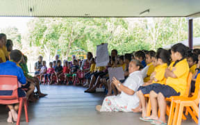 Niuean primary children in open air school assembly under cover of roof