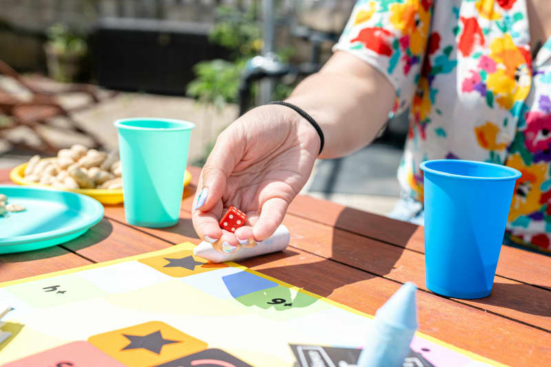 A person rolling the dice outdoors on a wooden table that has plates and cups.