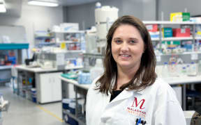 A profile photo of Kerry wearing a labcoat and standing in a lab. She is smiling with her shoulder length brown hair down. In the background are lab benches and shelves with coloured equipment.