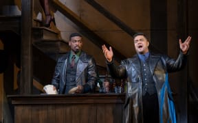 Pene stands on stage singing. He wears a large leather coat over a suit. Other actors glare at his character as he sings grandiosely.