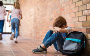 Little boy sitting alone on floor after suffering an act of bullying while children run in the background.