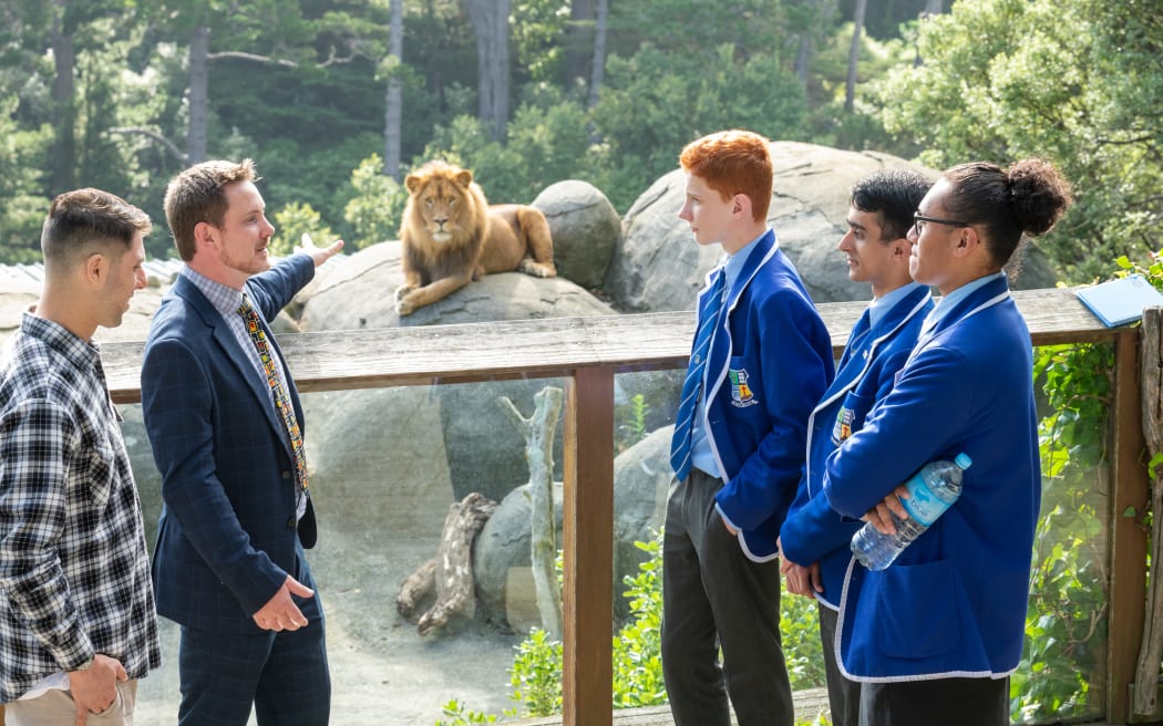 A man stands next to a zoo exhibit pointing at a lion while three high school students in blazers look on, and a man in a flannel shirt stands by.