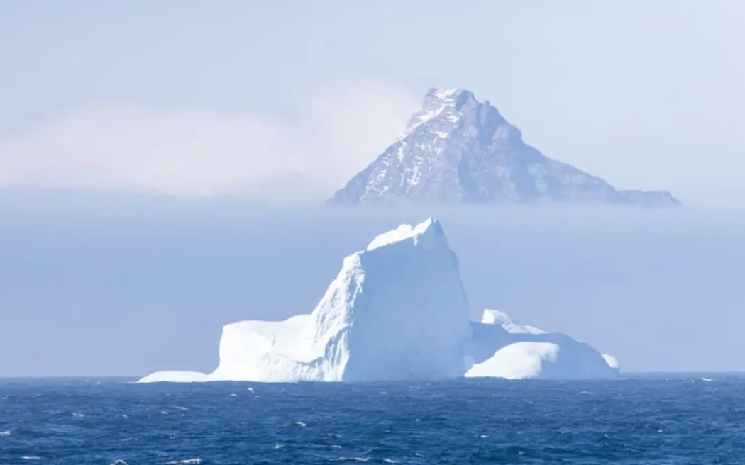 A pointy rocky mountaintop peeks out from behind a layer of thick fog. In the foreground is a large iceberg and dark choppy ocean.