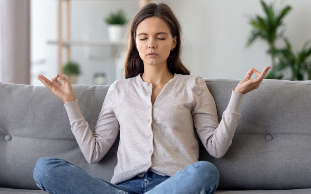 Peaceful young woman sitting in lotus pose on sofa in living room, meditating, attractive girl with closed eyes practicing yoga at home, stress relief concept, healthy good habit