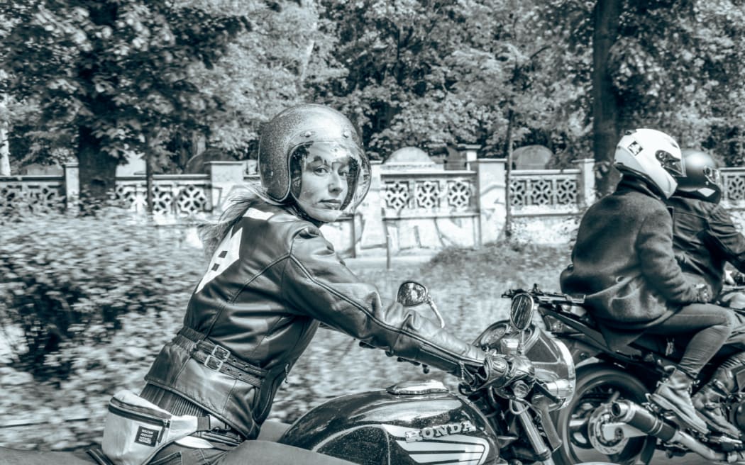A woman on a motorbike looks at the photographer with her hair flying out behind her.