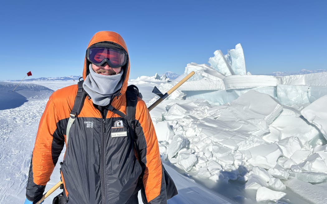 A man wearing black-and-orange Antarctica New Zealand-branded jacket with the hood up, a grey snood, a backpack and ski goggles stands on the ice smiling at the camera. He is carrying a long pole. Behind him are jumbles of ice slabs and a flag in the flat white expanse of ice beneath a bluebird sky.