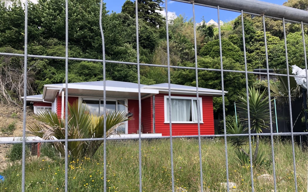 Behind a wire fence there is a small red house with an overgrown lawn around it. The left side of the house has been hit by a landslide. Behind the house are tall trees.