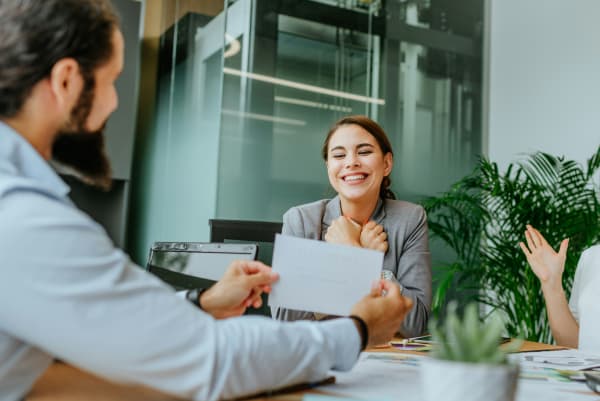 A woman smiles while sitting among colleagues, with one showing a card up to her.