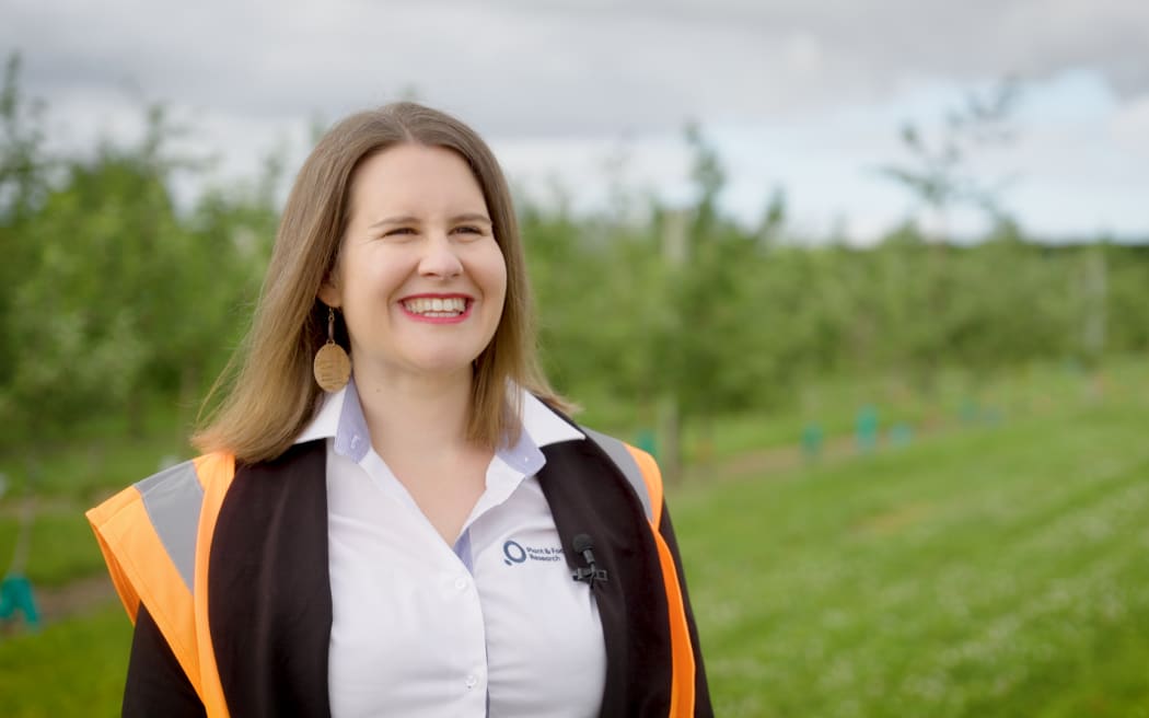 Emma Brown stands in an orchard. She has a big smile on her face. She is wearing a shirt, high vis, and big round bronze earings.