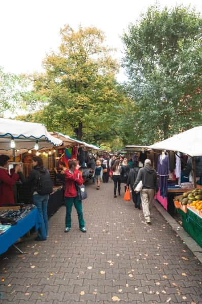 A group of people browse market stalls on a paved street with large trees in the background.
