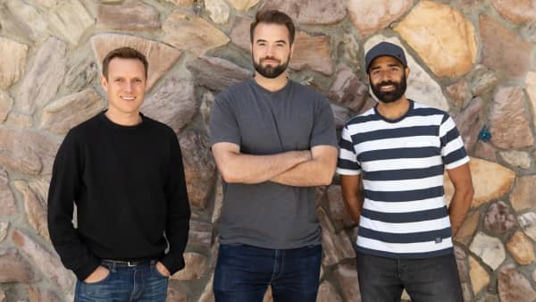 Three men in t-shirts stand against a wall.