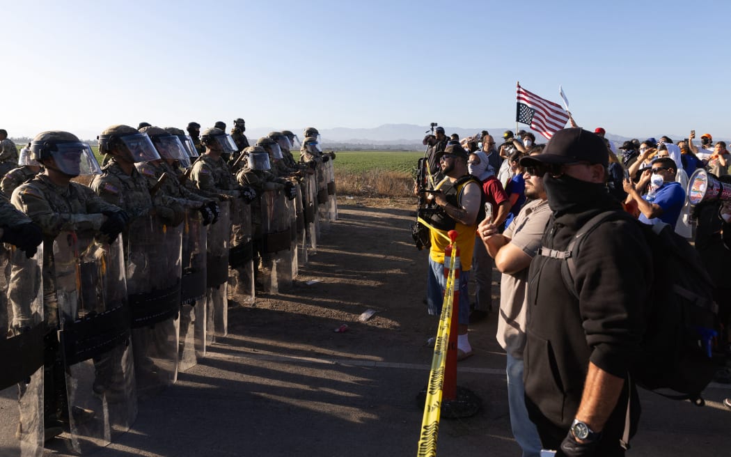 California National Guard troops face off with protestors during a Federal Immigration raid on Glass House Farms in Camarillo, California, July 10, 2025. Federal immigration agents—backed by CBP, Border Patrol units, National Guard troops, helicopters, and military style vehicles—raided Glass House Farms in Camarillo early July 10, 2025, targeting state licensed cannabis and other crop operations, detaining between a dozen and 100 farmworkers, and deploying smoke or tear gas canisters and rubber bullets to disperse a crowd of several hundred protesters on Laguna Road. (Photo by BLAKE FAGAN / AFP)