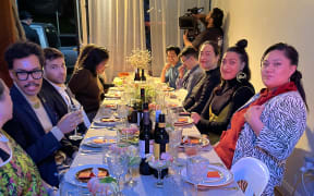 A photo of the dinner party in action. Guests are dressed up in festive 70s attire and seated on either side of a long table.