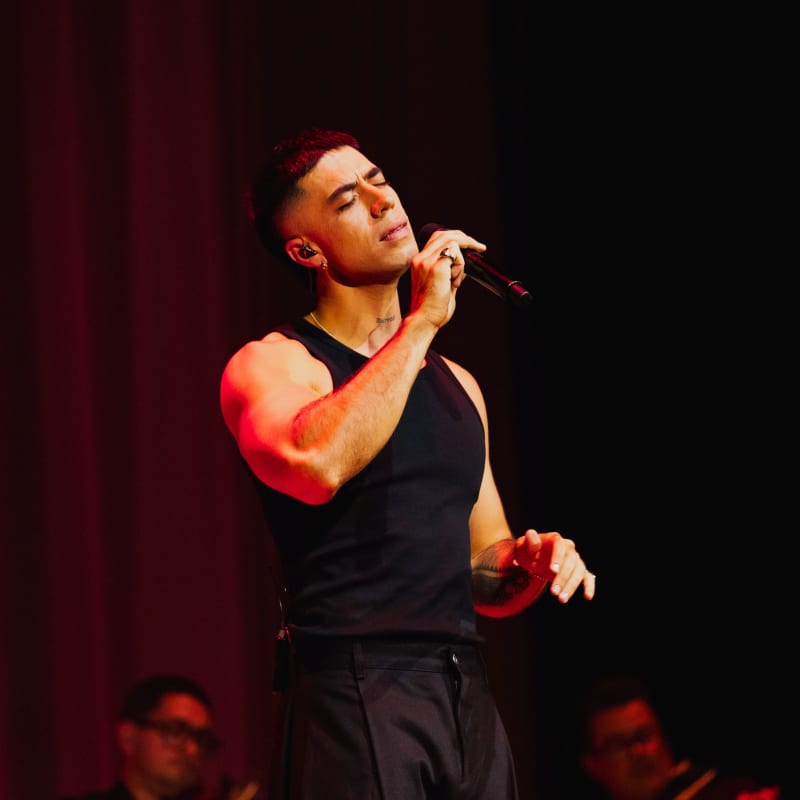 TEEKS, a muscular man in a black singlet and black jeans, sings into a microhpne with members of an orchestra and a red velvet curtain behind him.