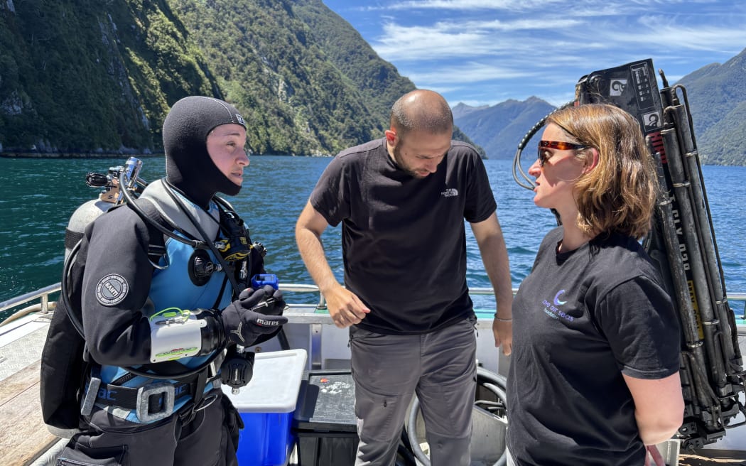 Three people onboard the deck of a boat. The person on the left is wearing a hooded wetsuit and full SCUBA gear and looking the woman on the right, who has on a dark t-shirt and sunglasses. The man in the middle, also wearing a t-shirt, is looking down. Behind the trio is blue ocean and steep-sided bushclad slopes.