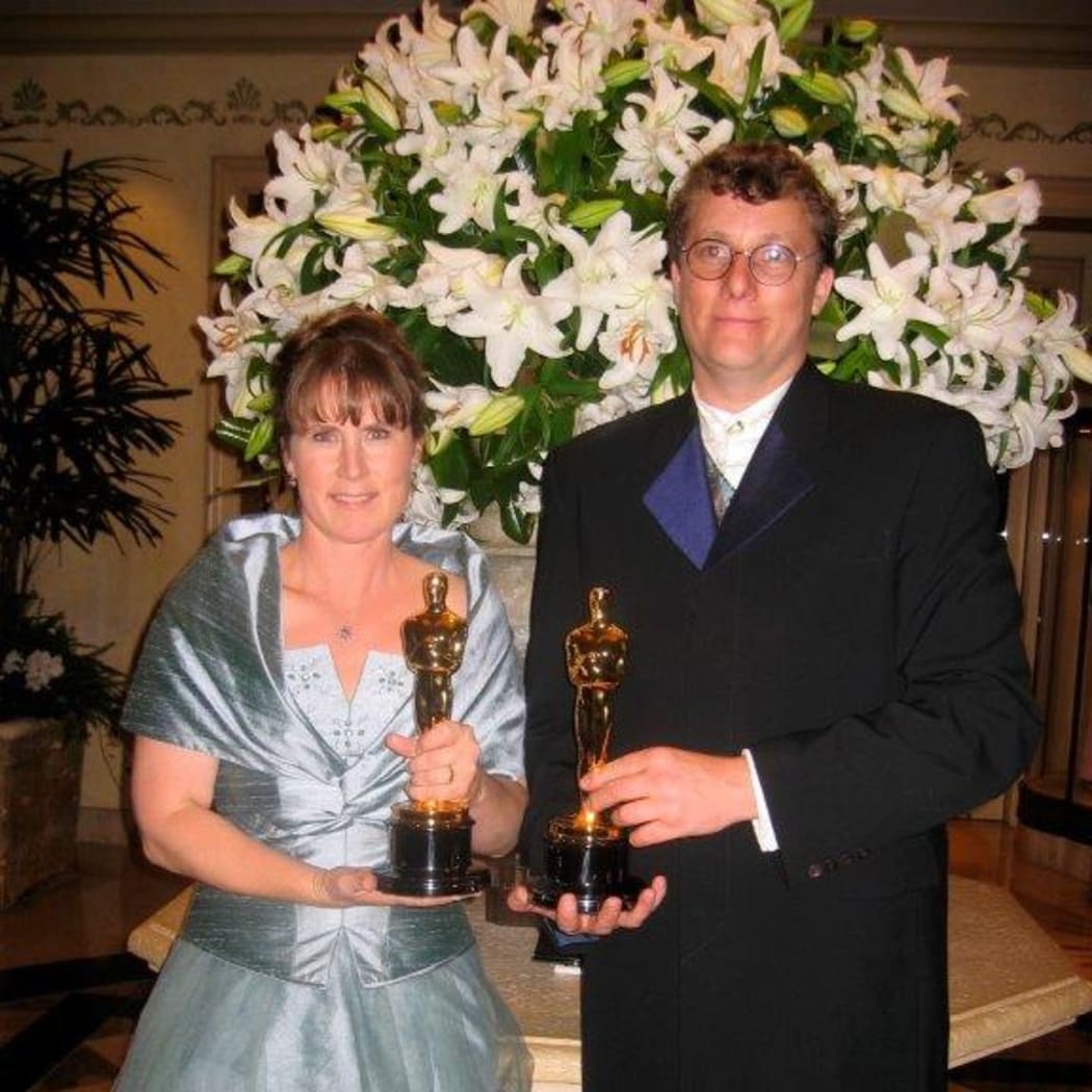 Tania and Richard stand side by side holding Oscars. They are both in fancy dress, Tania wearing a silvery blue ball gown and Richard wearing a black suit with satiny lapels.