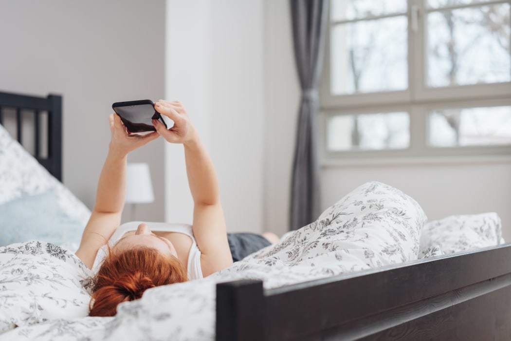 Young woman spending a relaxing day at home lying on her back on the bed reading a message on her mobile phone
