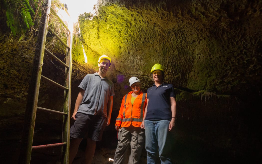 Three people wearing hard hats stand on a wooden platform in a cave, next to a ladder that ascends to a bright hole, where light enters and illuminates the cave ceiling.