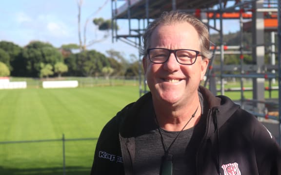 David smiles broadly at the camera. He is wearing a black jacket with a club shield logo on it. Behind him is a green football field and some scaffolding.