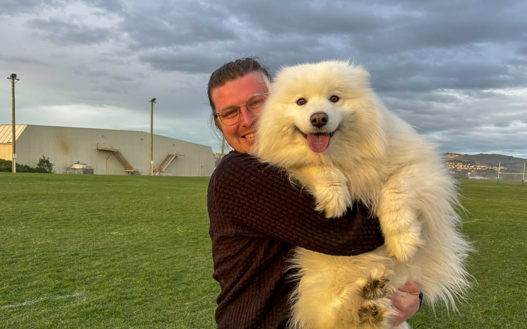 A man in a brown sweater smiles broadly as he lifts up his large fluffy white dog, a samoyed. They are both looking at the camera and standing in a grassy field in front of a large building.