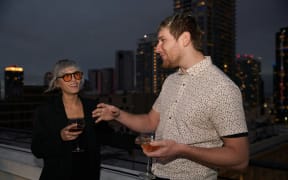 Two people holding wine glasses, standing on a building rooftop in a city.