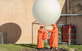 Two figures wearing long orange coats with orange hoods hold a large white balloon in front of a concrete wall and a collection of gas canisters.