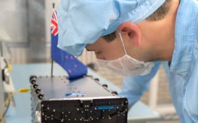 Ben Taylor is wearing a blue clean suit, including a hair net and facemask. He is building a satelite on a lab bench. The satelite has a New Zealand flag on it.