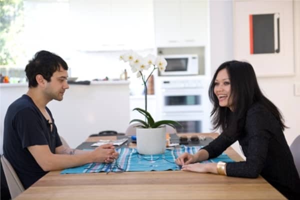 Kody Nielson and Bic Runga, two young people with blac hair and black tops, face each other across a table on which sits a potted lily.