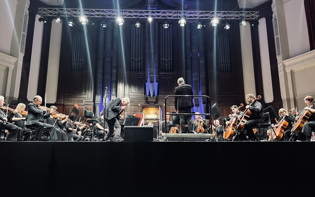 Ninety musicians sit in chairs in a semi-circle on a large stage. There are bright stage lights bearing down upon them. A conductor stands in the centre with his back to the camera. Shayne Carter is to the side, dancing and holding a microphone.