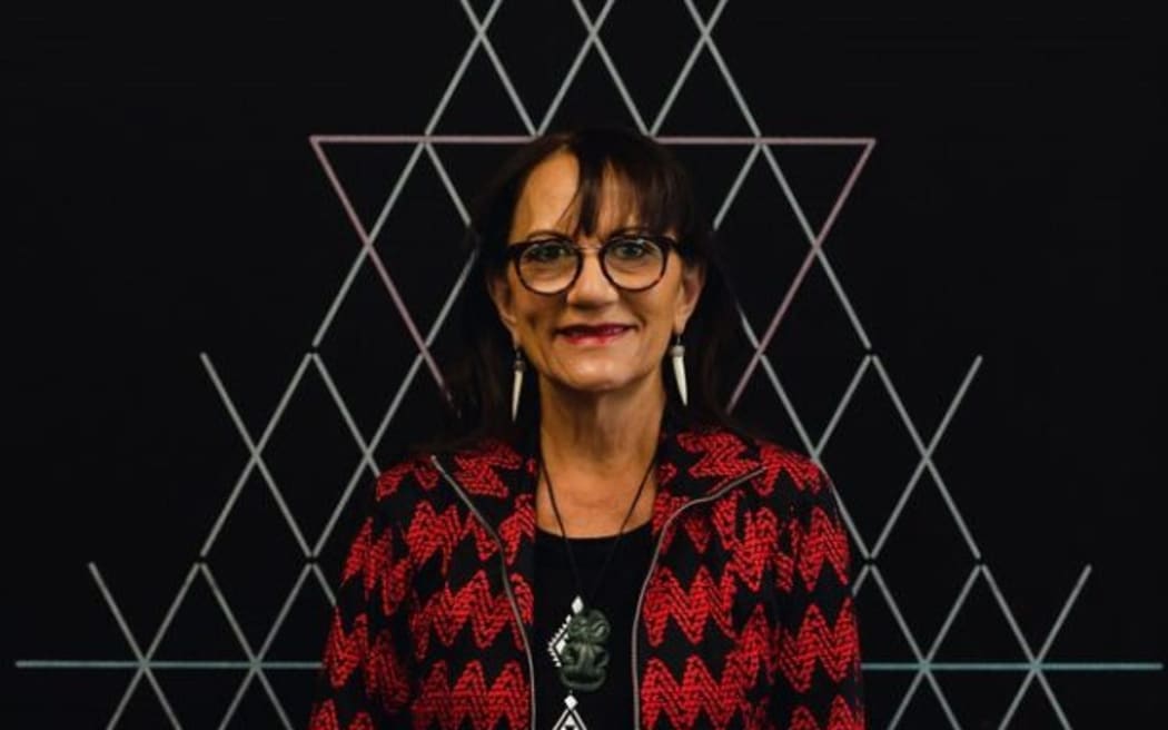 Te Puea smiles at the camera. She stands in front of a black wall designed with white tukutuku panelling.