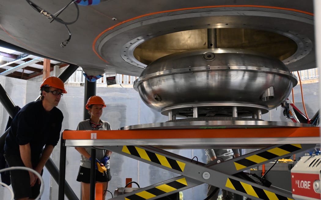 Two people wearing orange hard hats looking at a dinner-table-sized metal circular object on a pallet jack.