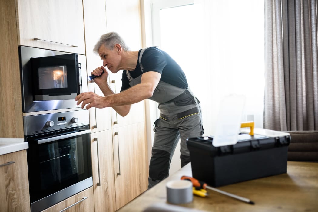 Aged repairman in uniform working, examining broken microwave in the kitchen using flashlight. Repair service concept. Selective focus.