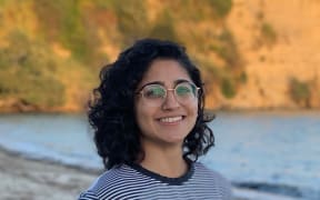 Sachi stands on a beach smiling at the camera. It is evening, and the rocky shoreline behind her is lit up with golden light.