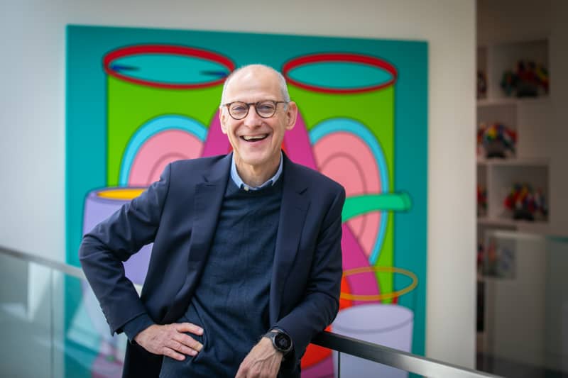 Dr Ezekiel Emanuel, a bald smiling man in a bule suit, leans with one hand on hip against a railing with a brightly coloured artwork behind him.