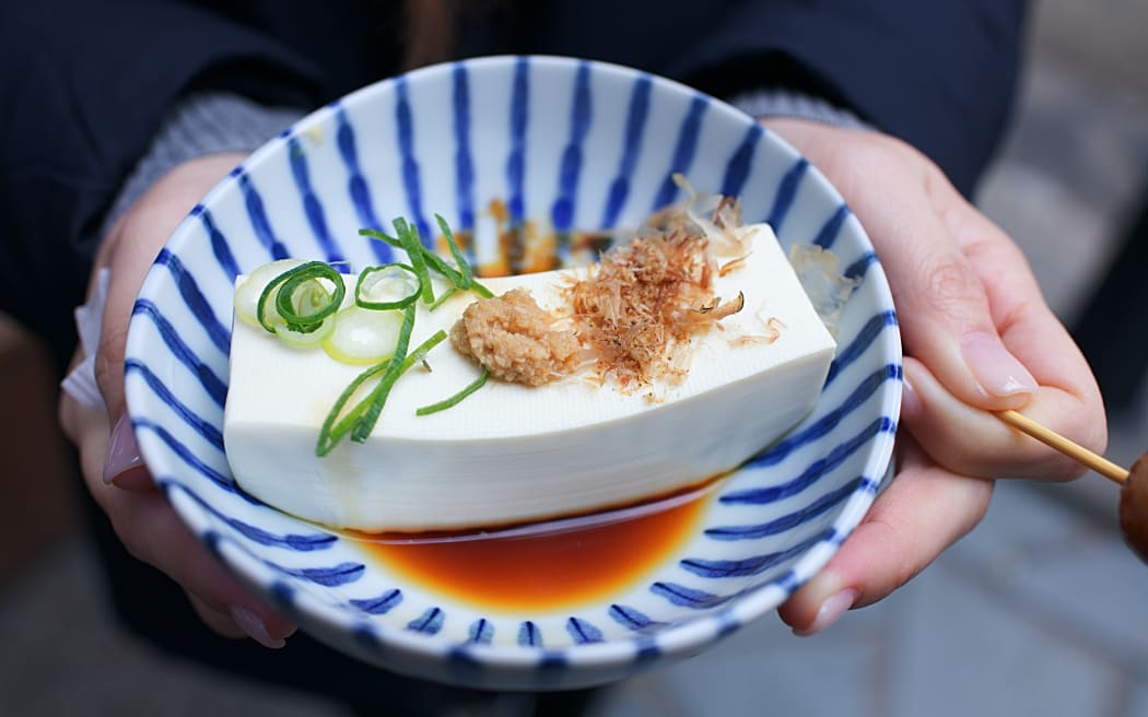 A blue striped bowl containing a piece of tofu bathed in soy sauce with spring onion garnish