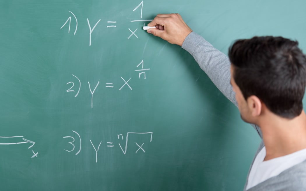 21205086 - male teacher with his back to the camera writing a mathematical formula on the blackboard in a classroom while teaching a class