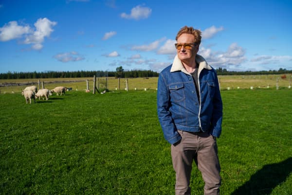 American TV host and comedian Conan O'Brien stands in a paddock with sheep in New Zealand in 2024.