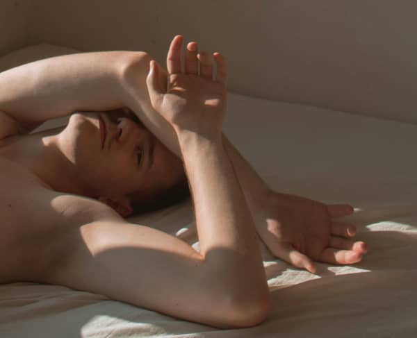 A pale-skinned man who is shirtless covers his face with this arms.