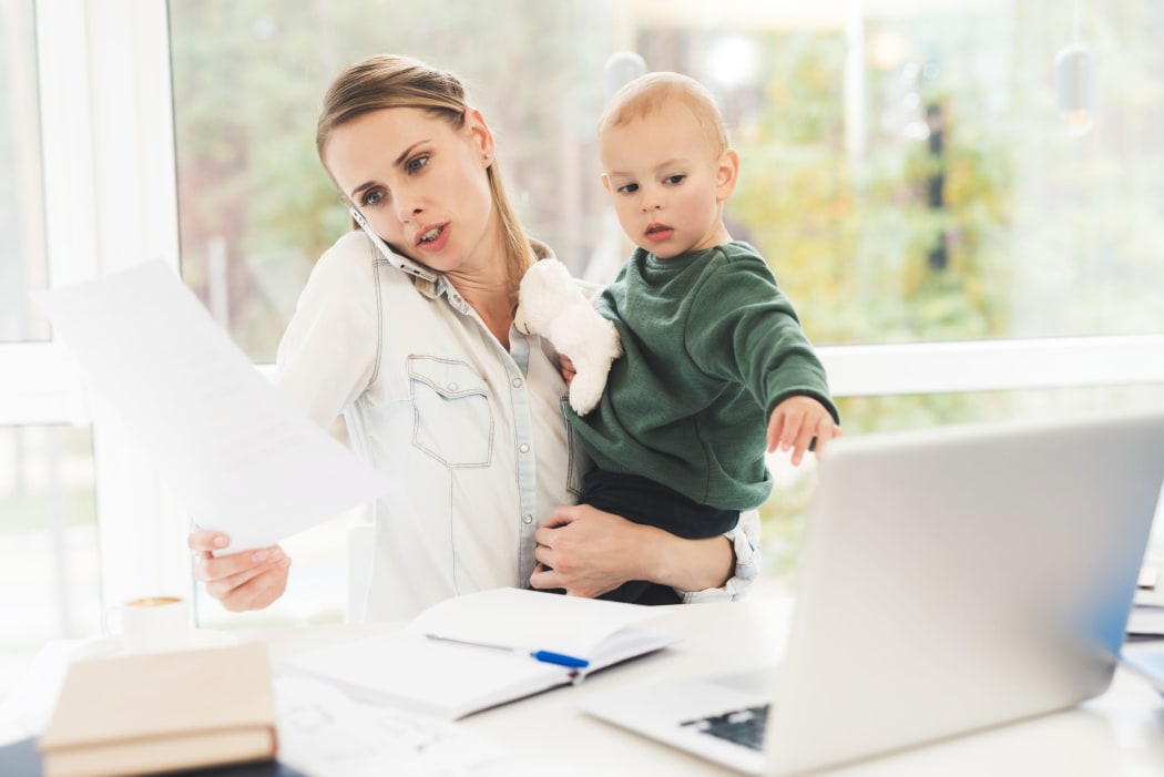 A photo of a woman who is working from home while she cares for a child at the same time. A woman with a child is sitting in a bright room.