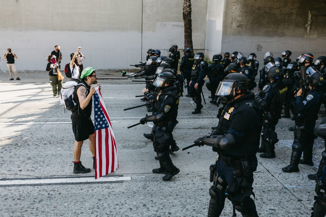 A protester stands holding a U.S. flag while facing a line of law enforcement officers during demonstrations in Los Angeles, California, on June 8, 2025. Following a series of aggressive federal immigration operations in Los Angeles, tensions escalated when Immigration and Customs Enforcement (ICE) agents conducted a high-profile raid on a Home Depot location. The raid sparked widespread protests across the city, where demonstrators decried the targeting of immigrant communities and the separation of families. Clashes soon broke out between protesters and federal agents, prompting U.S. President Donald Trump to deploy 2,000 National Guard troops to the city, a move harshly criticized by California officials, including the governor, as "purposefully inflammatory." (Photo by David Pashaee / Middle East Images via AFP)