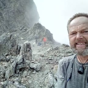 High up in Waiau Pass, Bruce pauses for a selfie in the background, two other walkers are climbing a steep, rocky ridge.