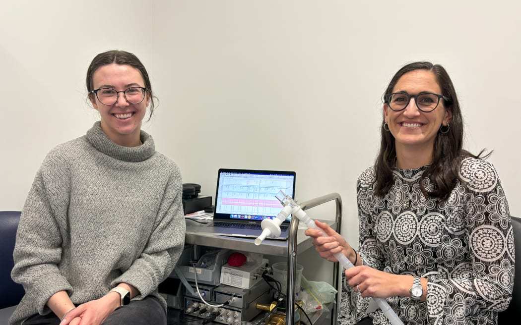 Two women with long dark hair and glasses smile and sit next to a laptop on a metal trolley in front of a white wall. On the tray underneath the laptop is a complex machine with a long tube, that the woman on the right is holding in her hands.