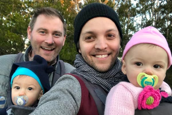 Two smiling men hold twin babies with dumbies in their mouths.